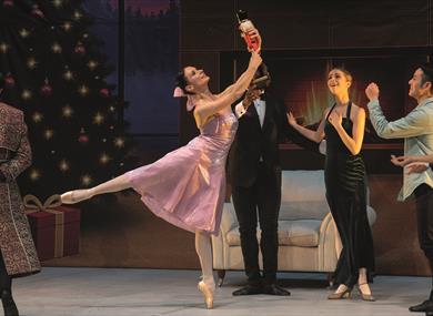 A female dancer wearing a pink satin party dress, her dark hair fastened in a bun with a pink bow, performs an arabesque. She's holding a Nutcracker d