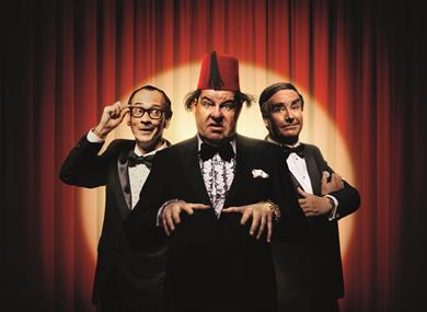 Three men stand together in front of a red curtain, lit with a golden spotlight. They all wear black tie. The man on the left wears glasses with thick