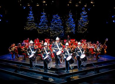The Band of HM Royal Marines stand on the Festival Theatre stage dressed in military full dress, facing out front. Five drummers stand at the front, w