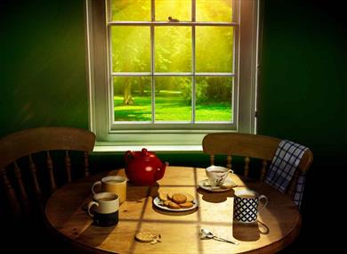 Three mugs of tea, a cup and saucer, a red teapot and a plate of digestive biscuits sit on a circular pine table. One slightly crumbled biscuit lies o