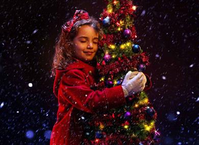 A young girl in a red winter coat hugs a small Christmas tree. She is smiling contentedly with her eyes closed, and has a red tartan bow in her hair. 