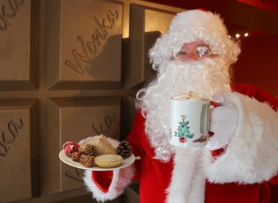 Santa stood in front of some giant chocolate bar doors holing a cup of hot chocolate and a plate of sweet treats, including a mince pie