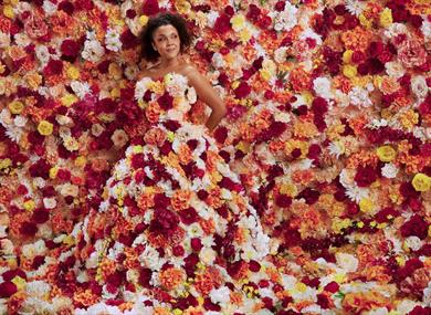 A young woman stands totally surrounded by flowers. They flood the background and cover her strapless, floor-length gown. The flowers are a mixture of
