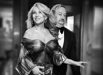 A black and white photo of a glamorous blonde woman in an off-the-shoulder pleated lurex dress and big gold hoop earrings, with a confident smile. Beh