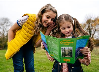 A child looking at a walk leaflet. She is stood in a green field, surrounded by trees