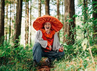 A person dressed up as a fungi crouching and smiling in the forest