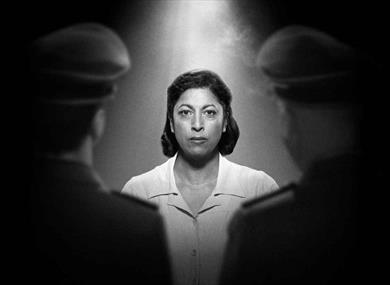 In a black and white photograph, a woman with dark shoulder-length hair stares resolutely at the camera, wearing a white top. A light illuminates her 