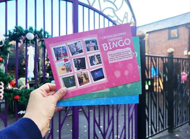 A hand holding a fan of bingo cards. In the bacground you can see some purple gates with a large Christmas wreath on them