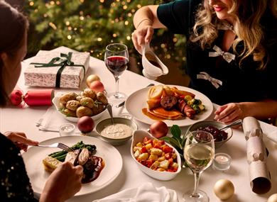 Guests enjoying a festive Christmas dinner with roast potatoes, seasonal vegetables, wine and all the trimmings beside a twinkling Christmas tree.