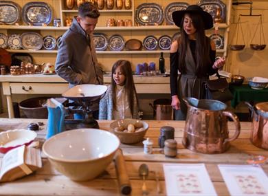 Three visitors stand around a wooden table in a historic kitchen or still room, surrounded by traditional cookware and kitchen tools at Uppark house a