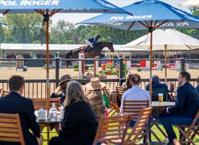 Royal Windsor Horse Show - people at tables under parasols watching showjumping