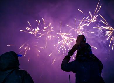 A family watching low bangs fireworks at Godstone Farm. The sky is purple and there are pretty fireworks. There is a lady and a man who has a child on