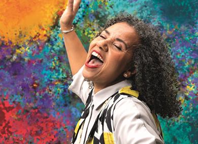 A woman sings joyfully, eyes closed and mouth open wide. Her head is passionately thrown back and her arm is raised. She has black and grey curly hair