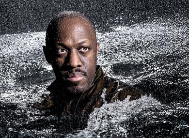 A man's head and shoulders rise from a sea of rough, churning water; rain is pouring down on him. He stares into the distance, his face illuminated by