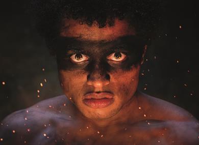A boy stares brutally straight into the camera with a straight face. There is messy black dirt over his eyes, like a mask, and he is topless with dirt