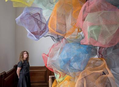 A women looks up a structure made from coloured mesh clouds that seem to float in the middle the stairwell of a wooden staircase.