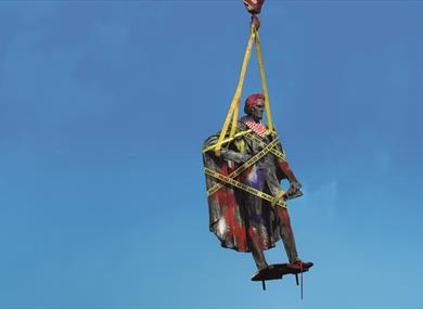 A traditional stone statue of a historic figure is suspended from an orange metal hook with yellow straps against a bright blue background. It is wrap
