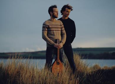 Two middle aged white men with dark hair and facial hair stand on the cliffs looking wistfully into the distance