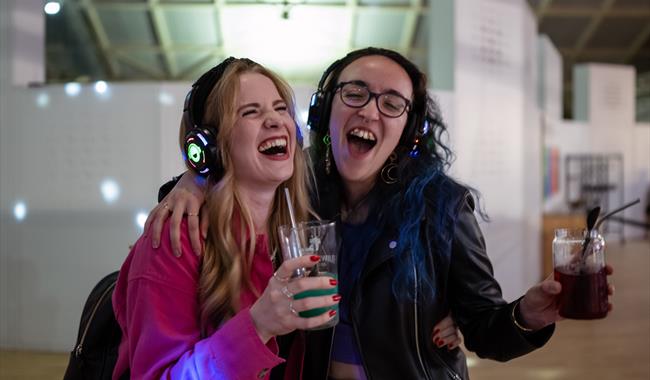 Two girls with headphones on singing and holding drinks