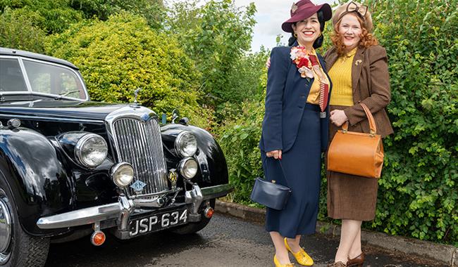 Two ladies dressed in 1940s fashion standing by a car from the 1940s