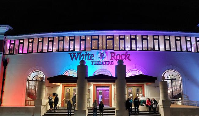 White Rock Theatre exterior lit at night with colourful lights and visitors gathered outside entrance.