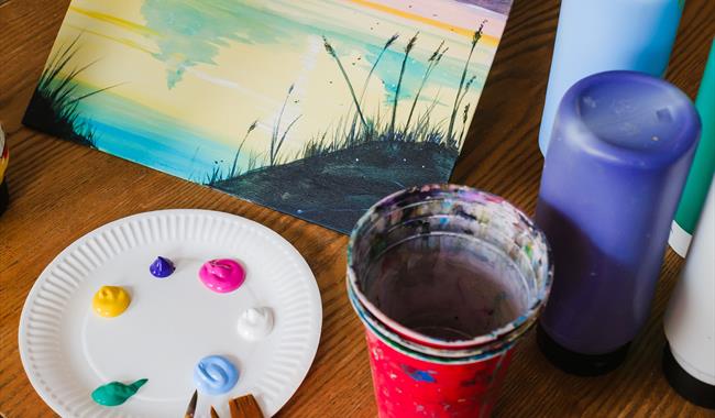 Painting class set up on a table including a tabletop easel with a finished landscape painting on canvas with red cup with paintbrushes neatly scatter