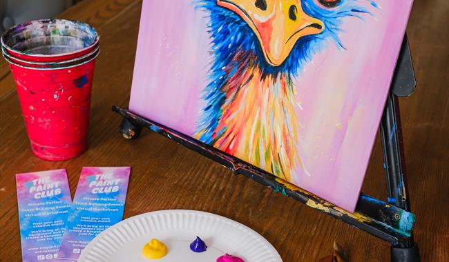 Painting class set up on a table including a tabletop easel with a finished landscape painting on canvas with red cup with paintbrushes neatly scatter