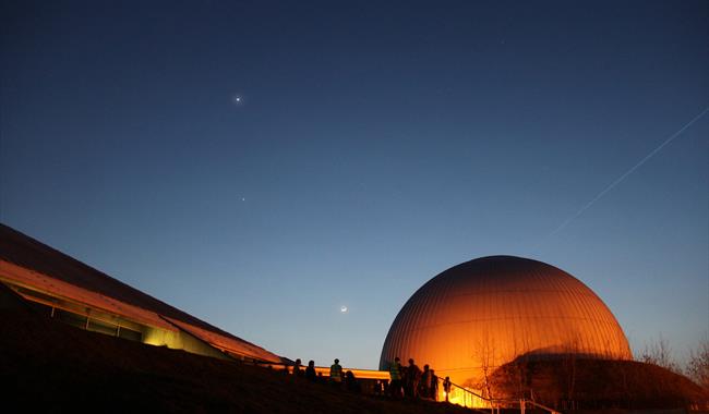 A photo outside the Planetarium. The Planetarium is glowing orange and there are a few stars in the night sky.