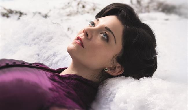 A young woman lies in the snow, looking up to the sky. She wears a rich purple silk dress with a high collar. She has brown hair and striking blue eye