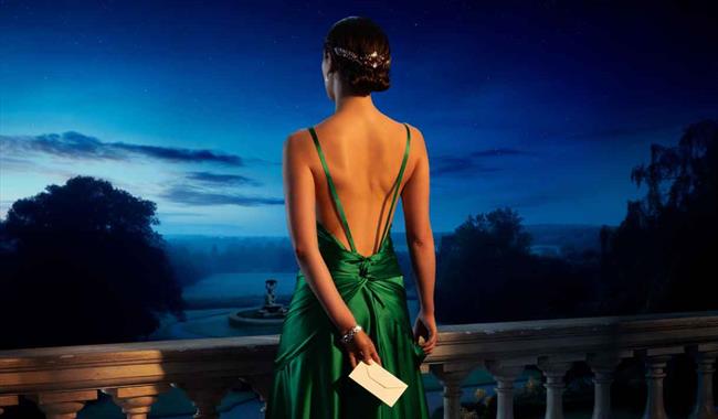 A young woman in a striking green backless dress holds a white envelope in one hand behind her back. She looks out at a garden and fountain from a bal
