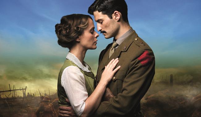 A couple embrace lovingly with solemn facial expressions. The man on the right wears military uniform and has dark hair and a moustache. The woman on
