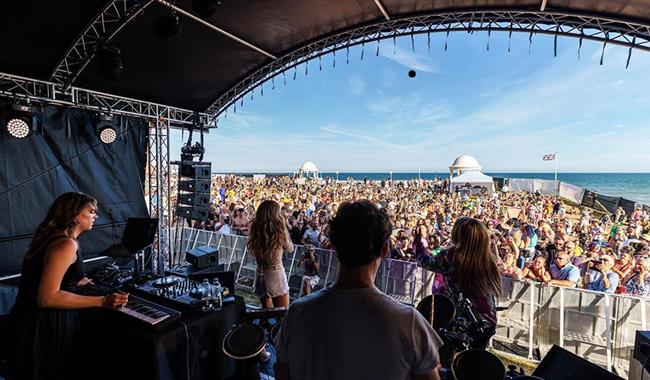 Photograph taken from within outdoor stage, look out at large crowd with the sea in the background.