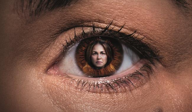 A close up of a woman's eye, within the pupil is an image of the same woman. She has dark brown hair and has a serious expression on her face.