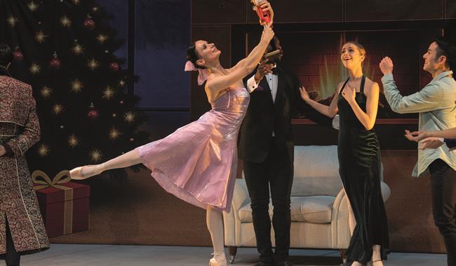 A female dancer wearing a pink satin party dress, her dark hair fastened in a bun with a pink bow, performs an arabesque. She's holding a Nutcracker d