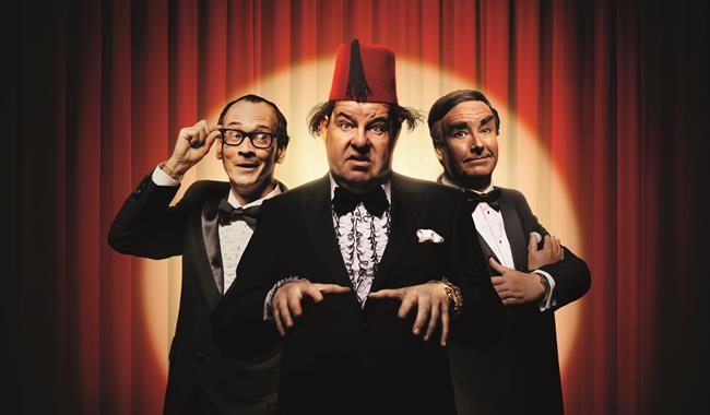Three men stand together in front of a red curtain, lit with a golden spotlight. They all wear black tie. The man on the left wears glasses with thick