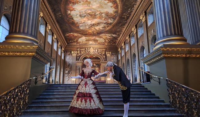 A man dressed in 18th-century attire is bowing to a woman in a lavish red and gold period gown on an ornate staircase in the Painted Hall.