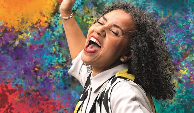 A woman sings joyfully, eyes closed and mouth open wide. Her head is passionately thrown back and her arm is raised. She has black and grey curly hair