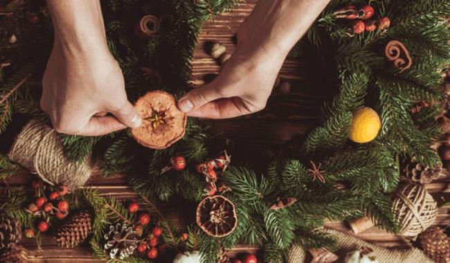 Hands decorating a Christmas wreath with dried orange slices, berries, pinecones and festive greenery on a wooden table.