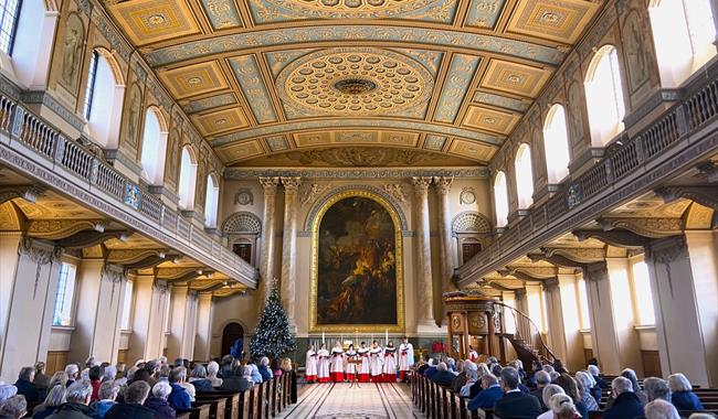 The Chapel of St Peter & St Paul, filled with people seated in pews, attending a Christmas carol concert. The choir, dressed in white robes with red a