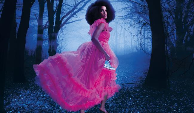 A woman in along, ruffled pink chiffon dress runs through a dark, misty woodland away from the camera. She is turning to look behind her, has an afro