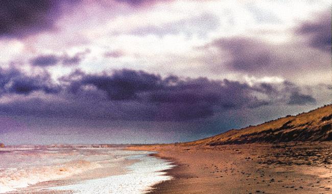 A moody watercolour image of a coastline under stormy skies. The sandy cove and dunes curve away into the distance, with waves breaking gently on the
