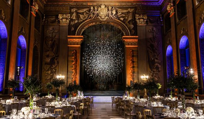 A rendering of the Painted Hall set up for a dining event with Coalescence in the background. The ceiling is lit up in orange and purple colours, and