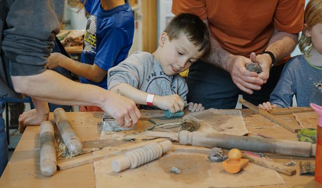 Young boy sitting at a table, stamping a pattern into a slab of clay. His parents are standing either side of him.