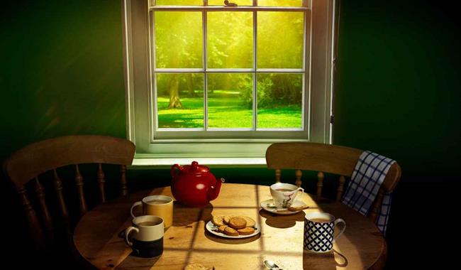 Three mugs of tea, a cup and saucer, a red teapot and a plate of digestive biscuits sit on a circular pine table. One slightly crumbled biscuit lies o