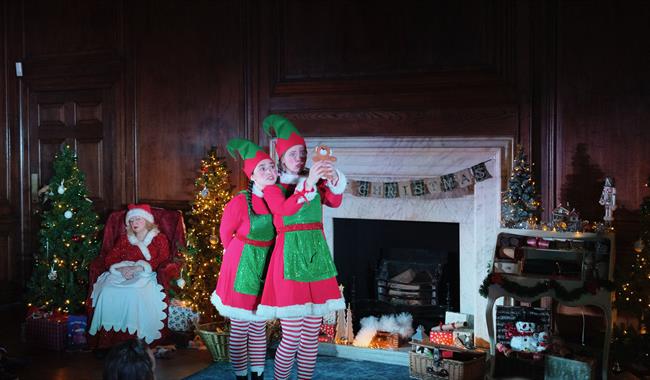 Two people dressed as Christmas Elves stand in front of a decorated fireplace. They are wearing green and red outfits with striped stockings and point