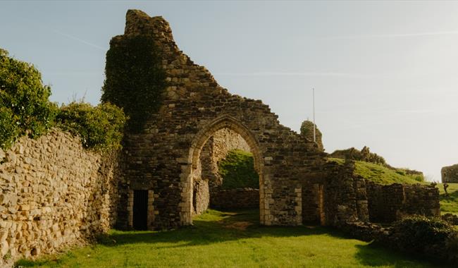 Stone archway and ivy-clad ruins of Hastings Castle in warm sunlight, with grassy grounds and hillside beyond.