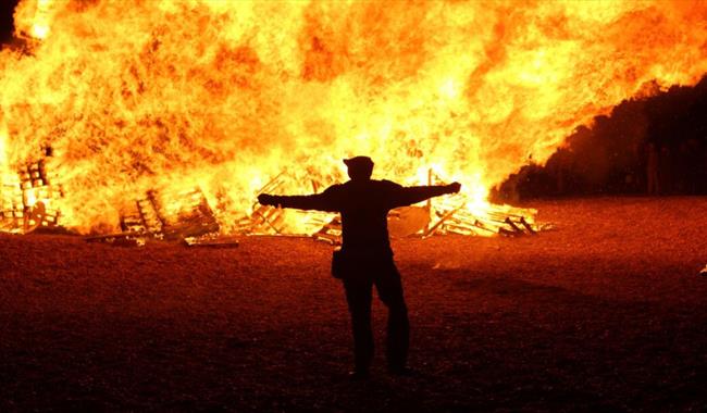Silhouette standing before a blazing bonfire, with towering flames lighting the night sky at a bonfire event.