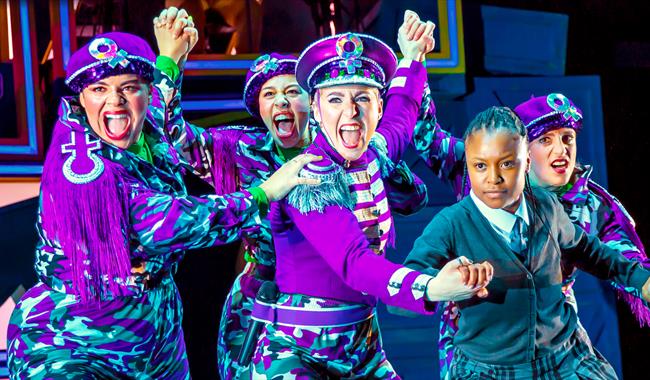 Four women in purple combat style trousers and purple military style jackets and hats are semi-crouched, facing out front, and clutching the hand of a