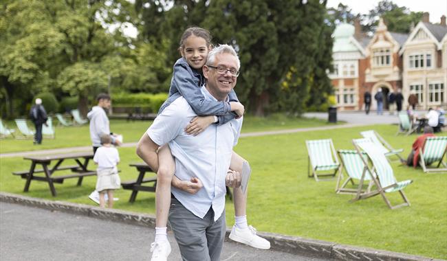 A father carries his daughter on his back as they walk past the Mansion at Bletchley Park. Both are smiling towards the camera.