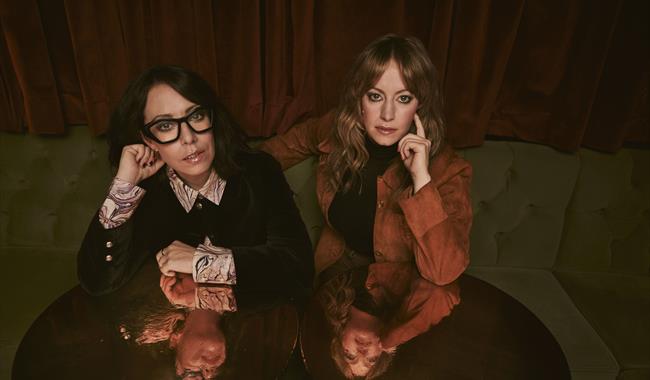Image: Flo and Joan sitting across sofa leaning on table